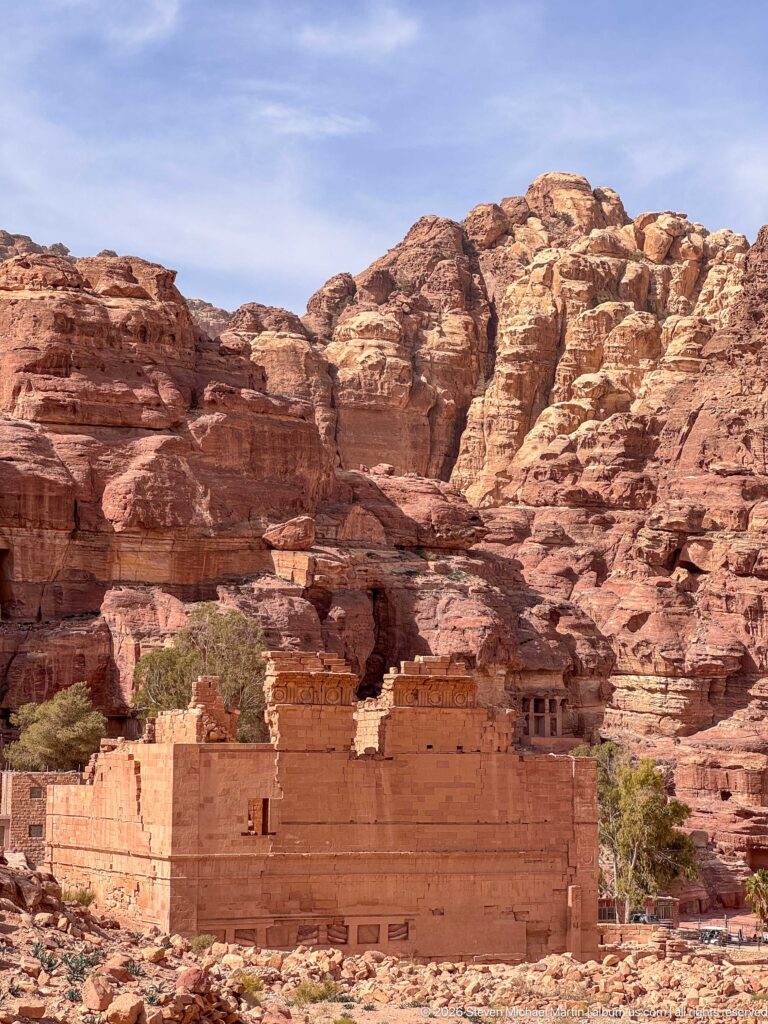 Long sandstone mass of Qasr al-Bint seen from a distance against layered Petra cliffs with rock-cut openings.