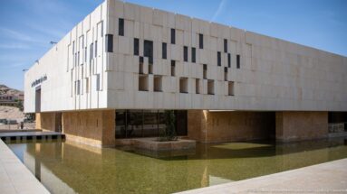 Long pale façade of the Petra Museum elevated above a shallow reflecting pool under a clear blue sky.