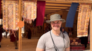 Smiling visitor in a brimmed hat stands among hanging patterned rugs and textiles inside a shaded Petra market stall.