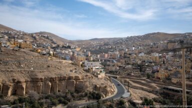 Hillside buildings of Wadi Musa cluster above a winding road amid dry, rocky slopes under a blue sky.