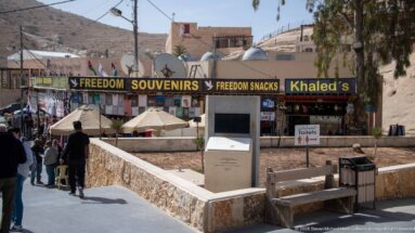 Visitors walk past souvenir and snack shops, benches, umbrellas, and signs near Petra’s entrance.