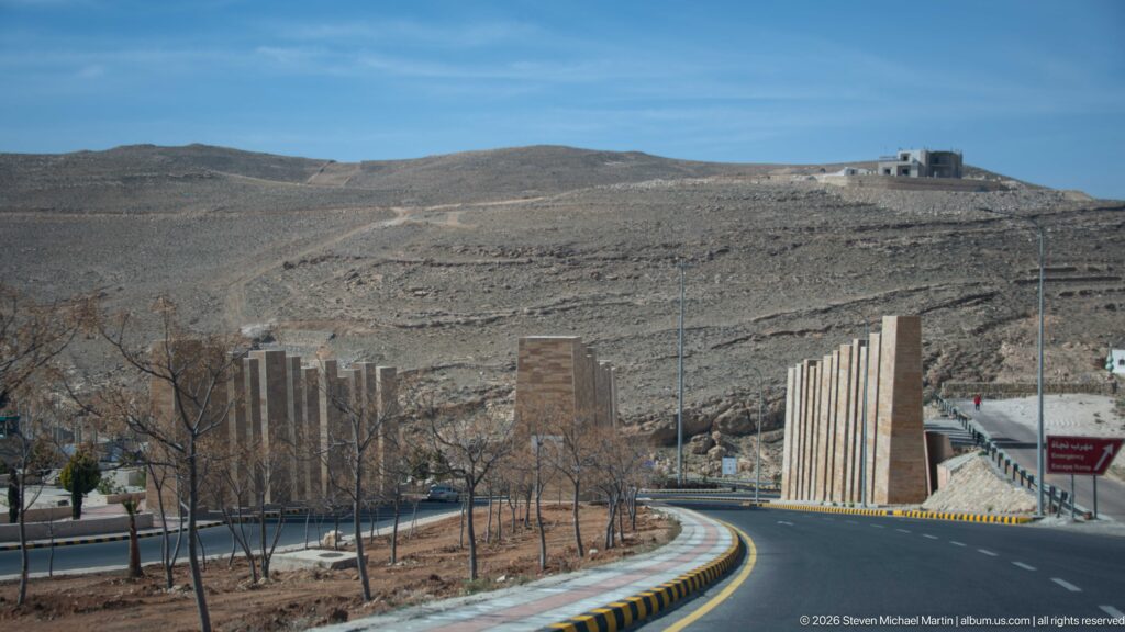 Curving modern road through barren Petra-region hills, lined with young trees and tall vertical roadside elements.