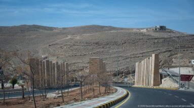 Curving modern road through barren Petra-region hills, lined with young trees and tall vertical roadside elements.