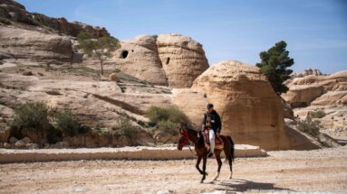 Rider on a brown horse crosses sandy ground before pale sandstone outcrops with rock-cut openings at Petra.