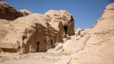 Rock-cut openings and weathered tomb façades spread across a pale sandstone outcrop near Petra’s entrance.