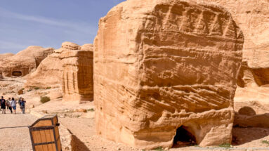 Large weathered freestanding sandstone block with a small opening at its base and visitors behind.