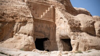 Rock-cut tomb façade with stepped crown and dark doorway carved into pale sandstone near Petra’s entrance.