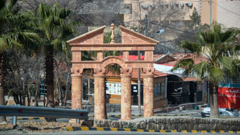 Decorative pink-and-tan gateway with columns, arch, and pediment beside a road and palm trees near Petra.