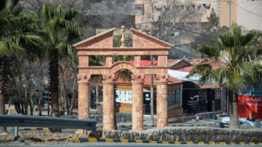 Decorative pink-and-tan gateway with columns, arch, and pediment beside a road and palm trees near Petra.