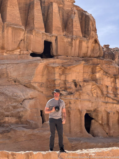 Visitor stands below Petra’s Obelisk Tomb, with four tapering monuments above and rock-cut chambers below.