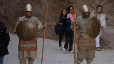 Two costumed performers hold long spears and round shields while tourists pass behind them at Petra.
