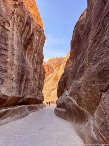 Broad bend of Petra’s Siq with high red sandstone walls and small visitors walking toward sunlit cliffs.