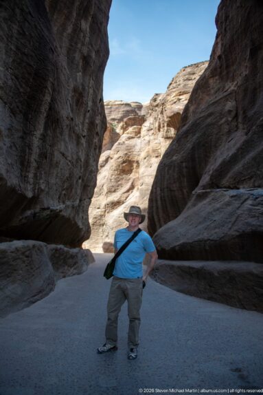 Visitor poses beneath dark overhanging sandstone walls in Petra’s Siq, with bright cliffs beyond.