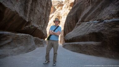 Visitor in blue shirt stands in Petra’s Siq between dark sandstone walls and a bright opening.