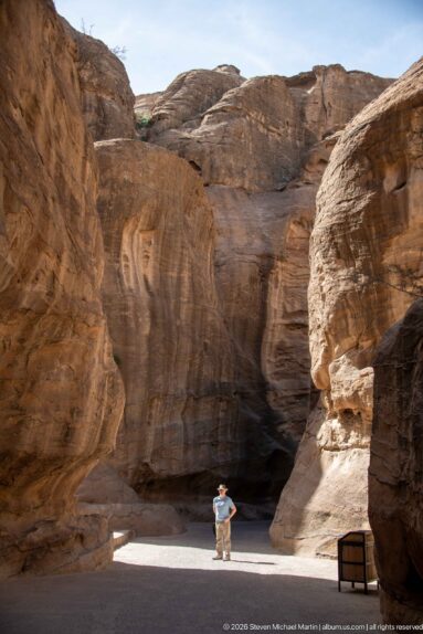 Visitor stands in a broad sandstone opening of Petra’s Siq before a deep shadowed recess.