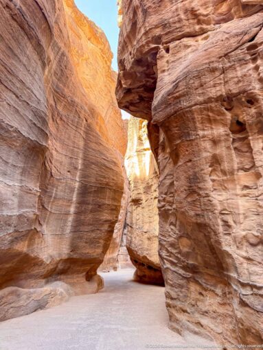 Layered red sandstone walls and a massive overhang frame a narrow opening in Petra’s Siq.