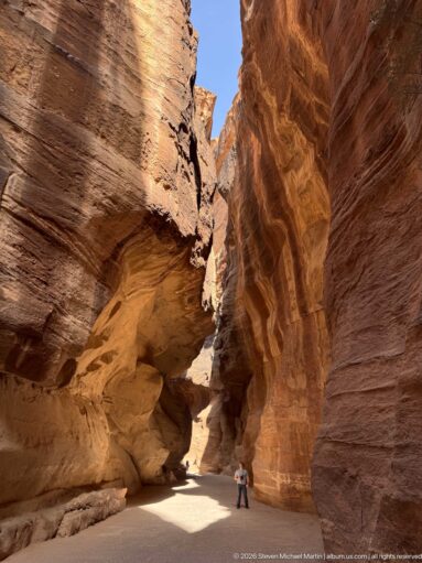 Layered sandstone overhangs rise above Petra’s Siq, with small visitors near a sunlit bend.