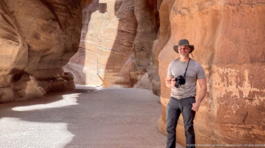 Visitor stands beside an orange sandstone wall in Petra’s Siq, with the curving canyon passage receding behind.