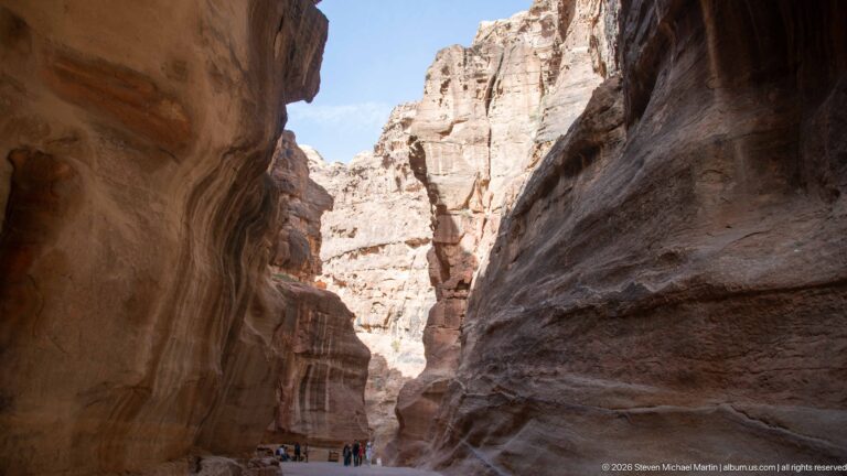 Visitors walk at the base of towering sandstone walls in a broader section of Petra’s Siq.