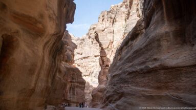 Visitors walk at the base of towering sandstone walls in a broader section of Petra’s Siq.