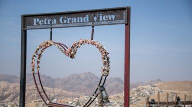 Flower-decorated heart frame beneath a Petra Grand View sign overlooking distant mountains and town.