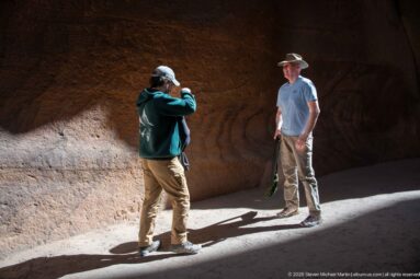 One visitor photographs another standing in bright sunlight against a dark sandstone wall in Petra’s Siq.