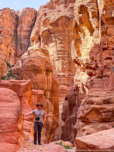 Visitor stands below red, white, and ocher banded sandstone cliffs with rounded cavities at Petra.