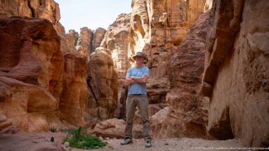 Visitor in sun hat stands on a sandy ledge among towering, weathered sandstone walls in Petra’s Siq.