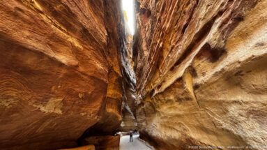 Towering banded sandstone walls of Petra’s Siq converge toward a narrow strip of sky above tiny visitors.