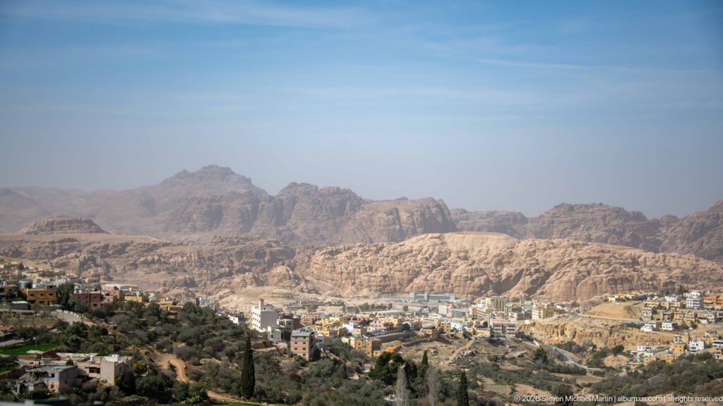 Rugged Petra mountains rise beyond Wadi Musa buildings and patches of vegetation under a pale blue sky.