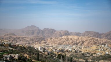 Rugged Petra mountains rise beyond Wadi Musa buildings and patches of vegetation under a pale blue sky.