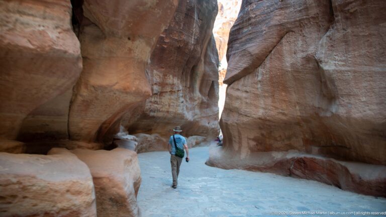 Visitor walks through a tall narrow corridor of Petra’s Siq between smoothly eroded sandstone walls.