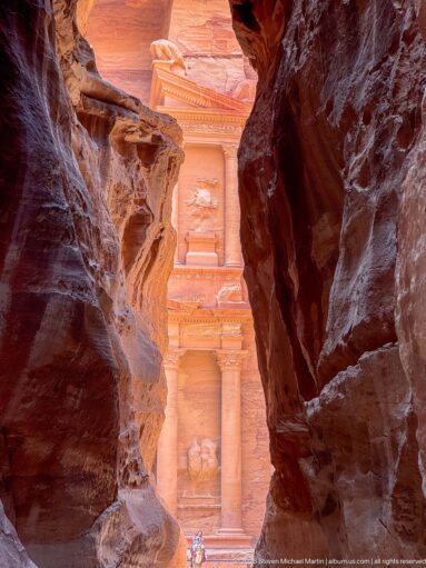 Partial view of Petra’s Treasury seen through a narrow opening between dark sandstone walls of the Siq.