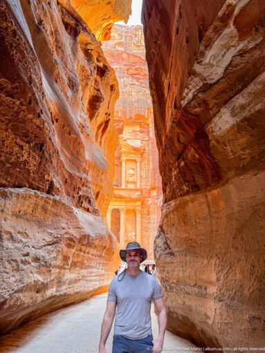 Visitor stands in Petra’s narrow Siq with the sunlit Treasury visible between curving sandstone walls behind him.