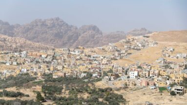 Panoramic Wadi Musa townscape below rugged Petra mountains, with dense buildings and darker cultivated slopes.