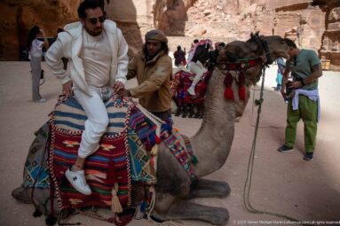 Visitor mounts a kneeling camel with colorful saddle blankets while a handler stands beside it at Petra.