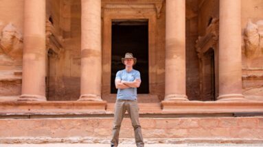 Single visitor stands centered between sandstone columns before the dark entrance of Petra’s Treasury.