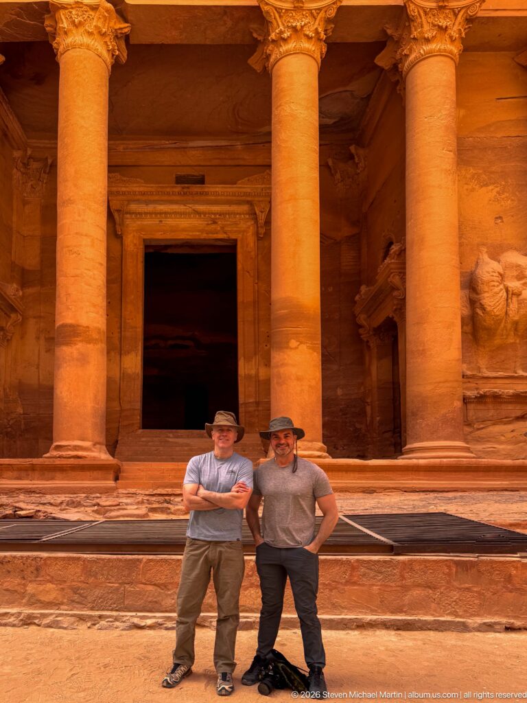 Two visitors stand beneath massive Treasury columns in front of the dark central doorway at Petra.