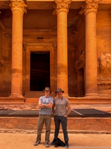 Two visitors stand beneath massive Treasury columns in front of the dark central doorway at Petra.
