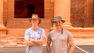 Two smiling visitors in hats pose before the steps, columns, and entrance of Petra’s Treasury.