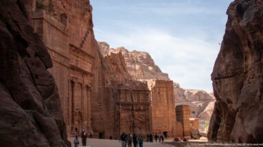 Visitors walk through a narrow Petra passage beside tall rock-cut façades receding toward distant mountains.
