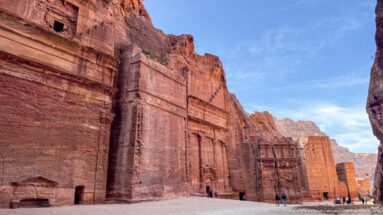 Monumental rock-cut façades recede along Petra’s sandstone wall with visitors walking at their base.