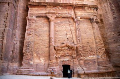 Columned rock-cut sandstone façade with central doorway and two visitors standing at the entrance.