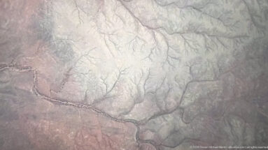Aerial view of pale desert terrain cut by branching dry channels and subtle ridges.
