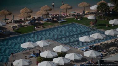 Elevated view of a patterned swimming pool with white umbrellas, lawns, thatched beach parasols, and calm blue water.