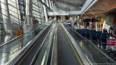 Long moving walkway rising through a glass-sided airport corridor with seating, shops, and overhead signs.