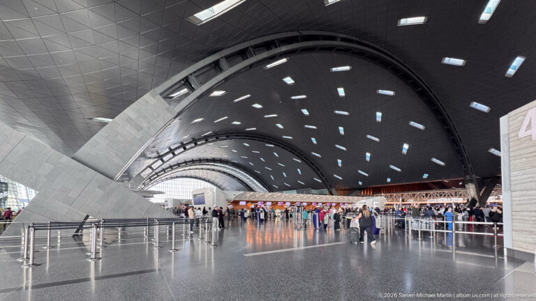 Wide airport departure hall with sweeping curved roof, rectangular skylights, massive angled supports, and distant passengers.
