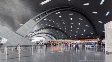 Wide airport departure hall with sweeping curved roof, rectangular skylights, massive angled supports, and distant passengers.