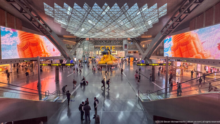 Wide airport concourse with giant digital screens, travelers, escalators, and the yellow Lamp Bear in the distance.