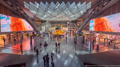 Wide airport concourse with giant digital screens, travelers, escalators, and the yellow Lamp Bear in the distance.